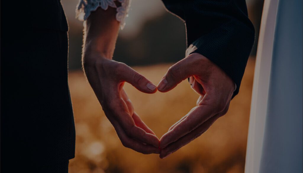 A close-up shot of a couple’s hands forming a heart shape against a golden hour background. The person on the left wears a white lace sleeve, while the person on the right is in a dark suit jacket.