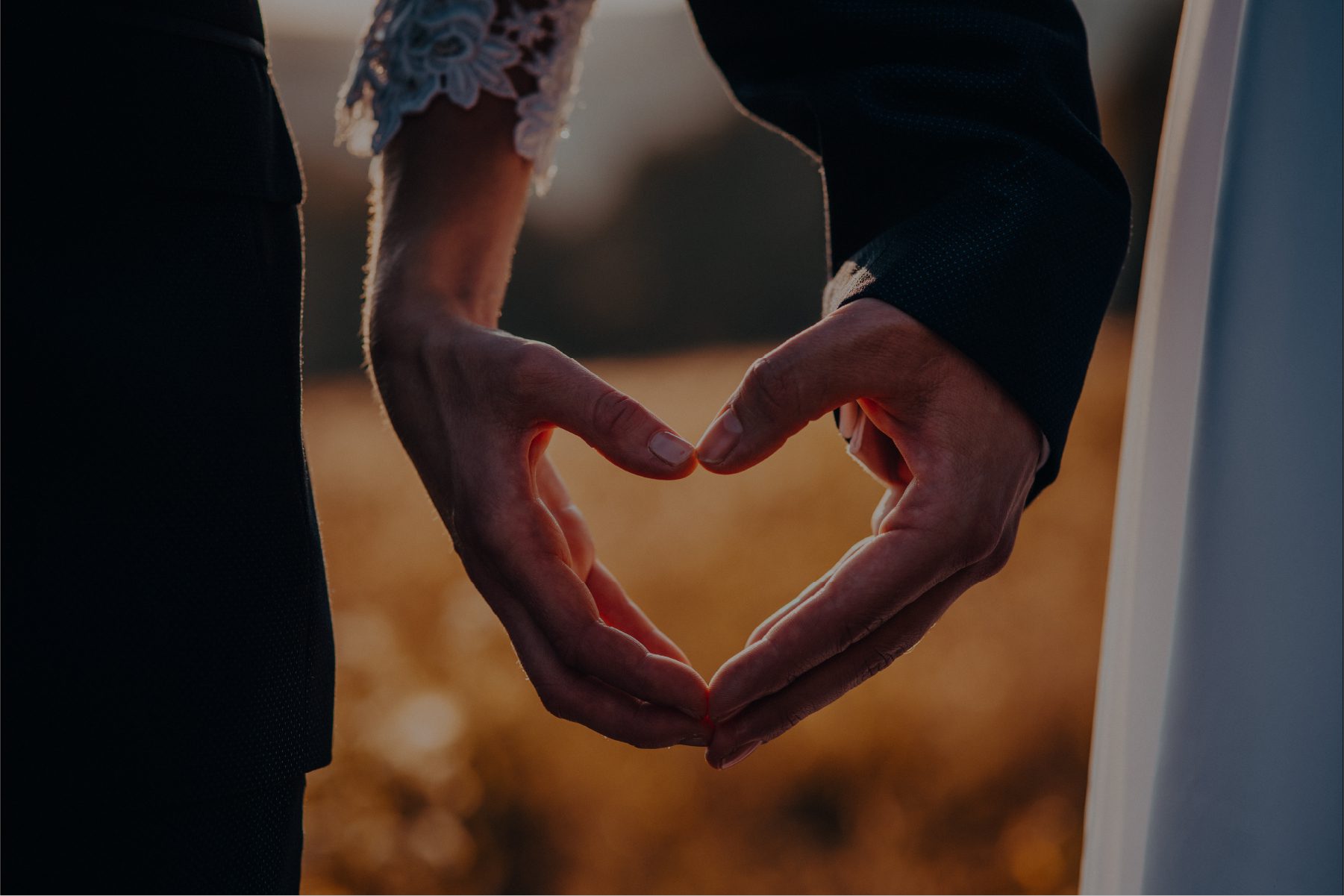 A close-up shot of a couple’s hands forming a heart shape against a golden hour background. The person on the left wears a white lace sleeve, while the person on the right is in a dark suit jacket.
