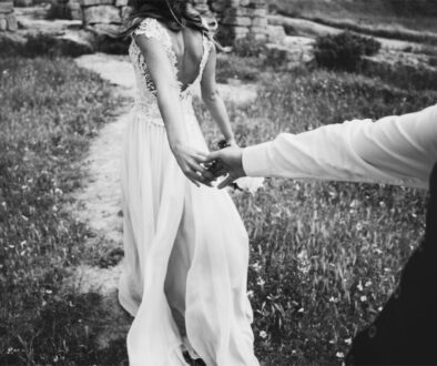 A bride in a white wedding dress leading her partner by the hand through a field.