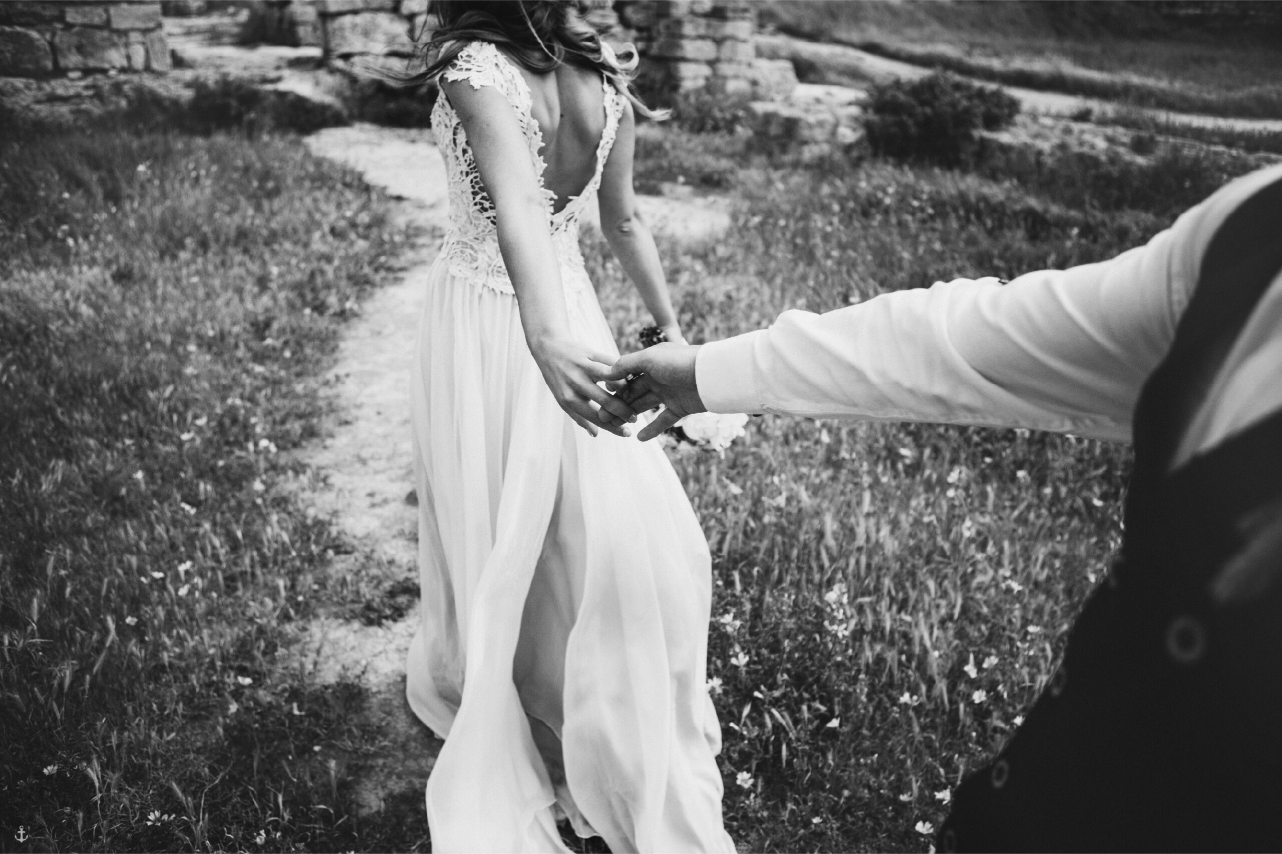 A bride in a white wedding dress leading her partner by the hand through a field.