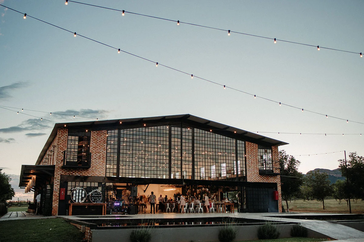 A modern-industrial brick wedding venue at dusk featuring a massive facade of black-framed glass grid windows glowing with warm light, string lights overhead, and a reflecting pool in the foreground.