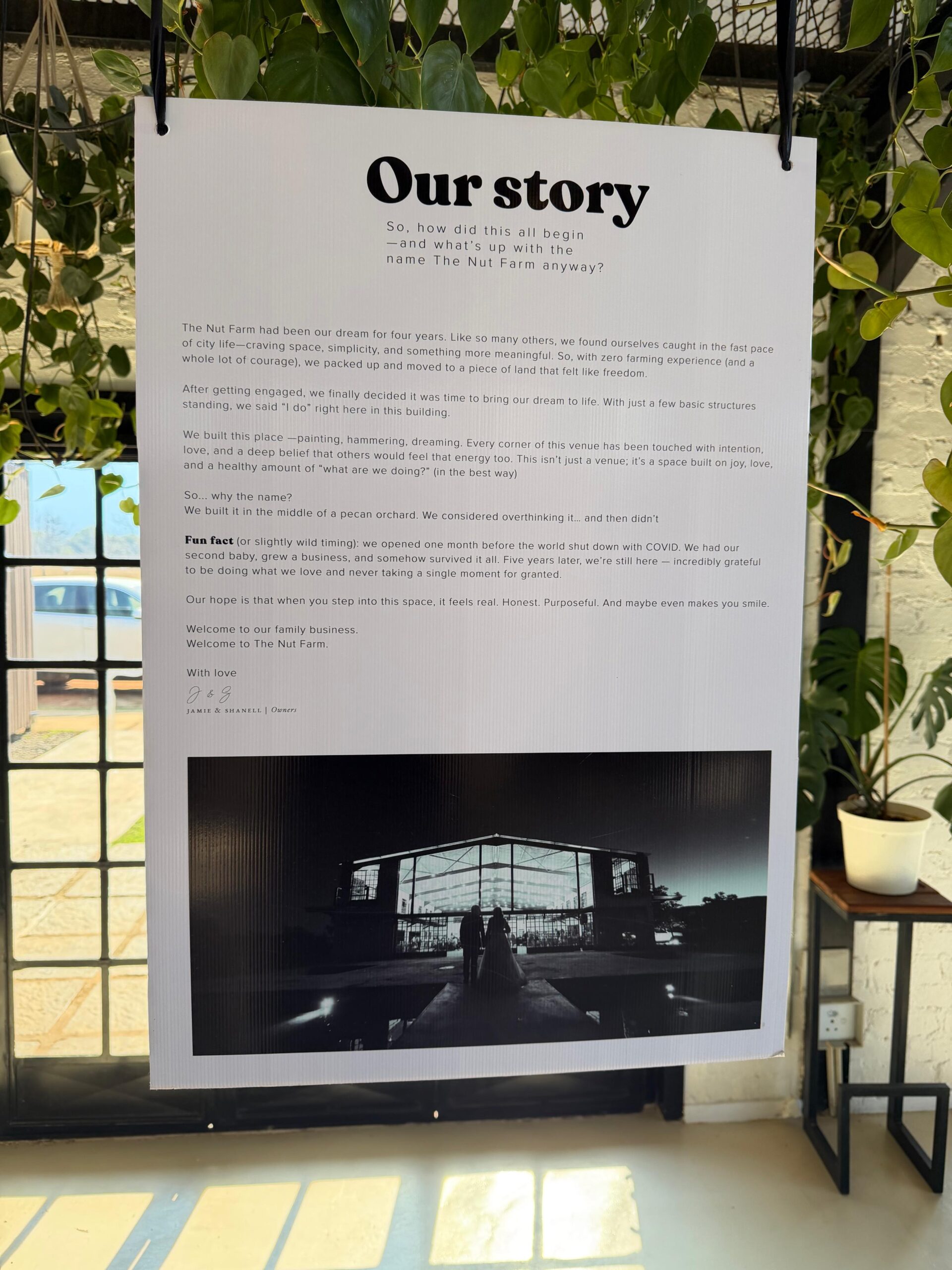 A hanging white sign titled "Our story" detailing the history of The Nut Farm wedding venue, featuring a printed black-and-white photograph of a bride and groom standing in front of a modern glass-and-steel greenhouse structure.